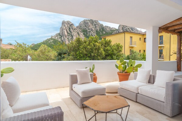 Un balcón con mobiliario de mimbre, plantas en macetas y vista a montañas rocosas.