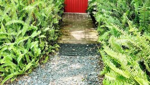 A red door with the number 115, surrounded by lush green ferns and palm trees. - Steps to Dining, Shopping, 3 Minute Walk to Beach (Folly Beach)