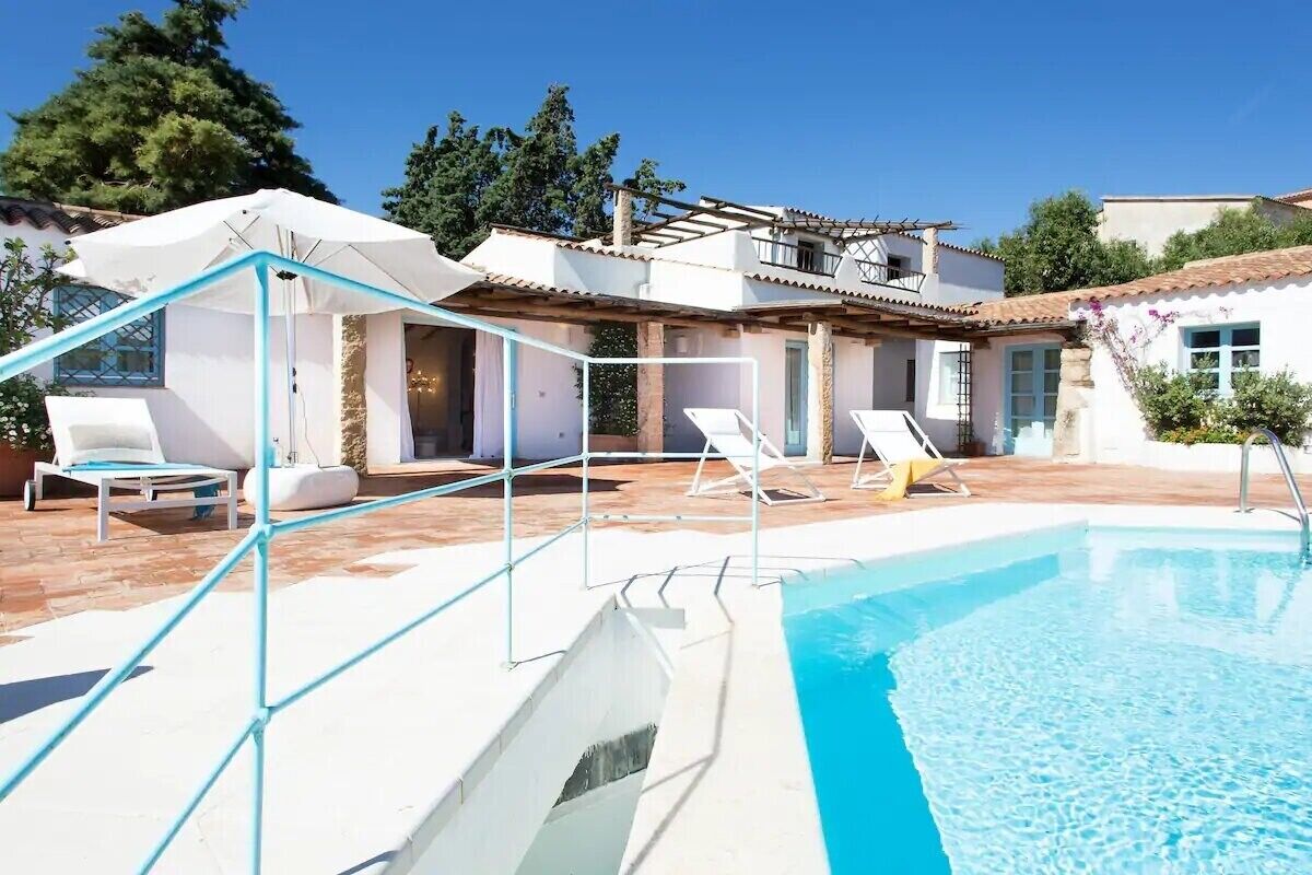 Un área junto a la piscina con sillones de descanso, un espacio de estar cubierto y un edificio con tejado de tejas.