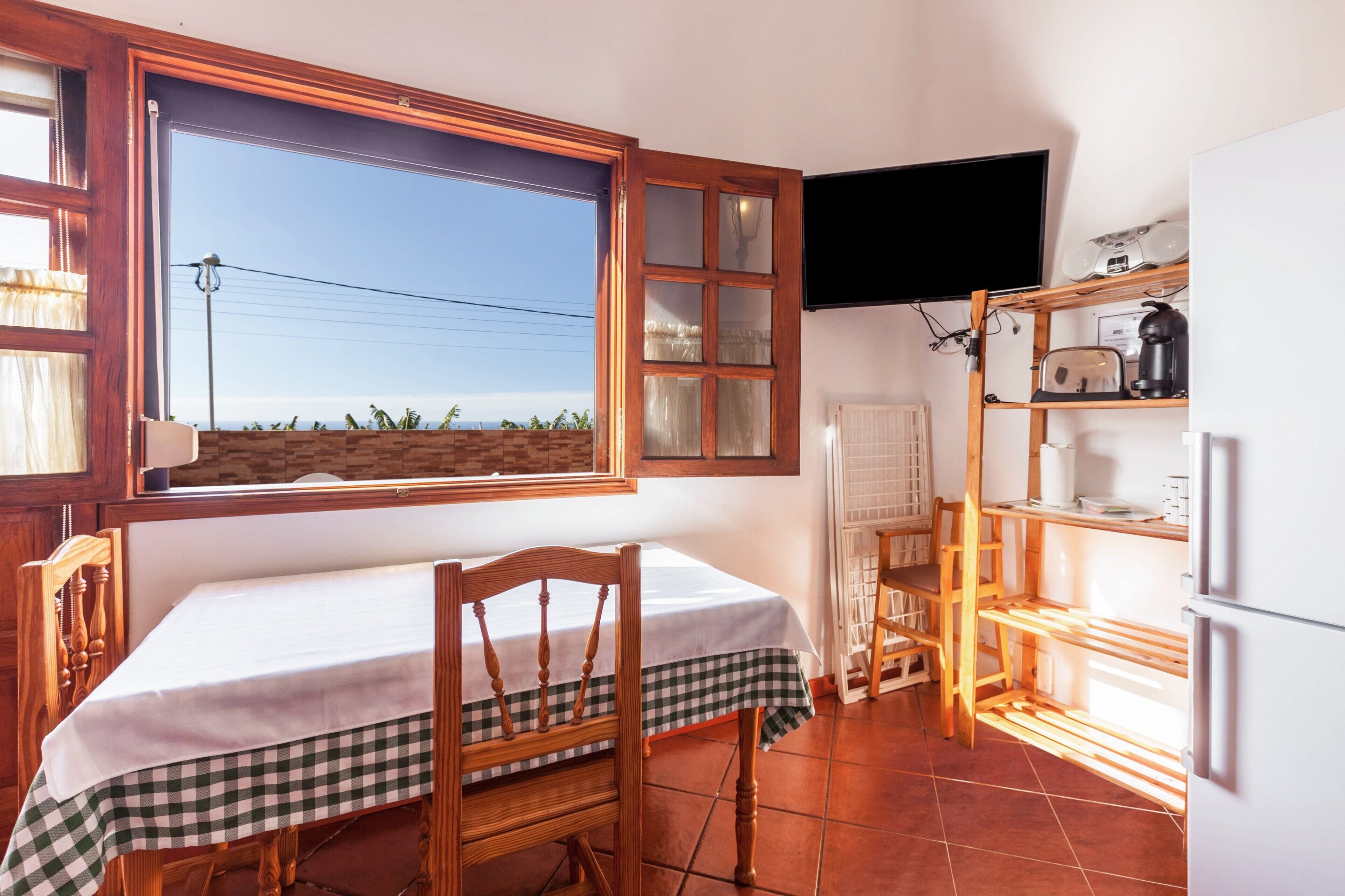Eine Küche mit Fenster, einem Tisch mit kariertem Tischläufer, einem Holzstuhl, einem an der Wand befestigten Fernseher und einem Kühlschrank.