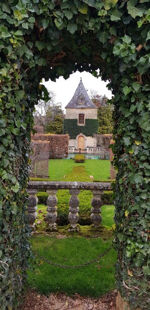 Un bâtiment en pierre surmonté d’un toit conique, entouré de haies bien entretenues et d’une pelouse parfaitement taillée.