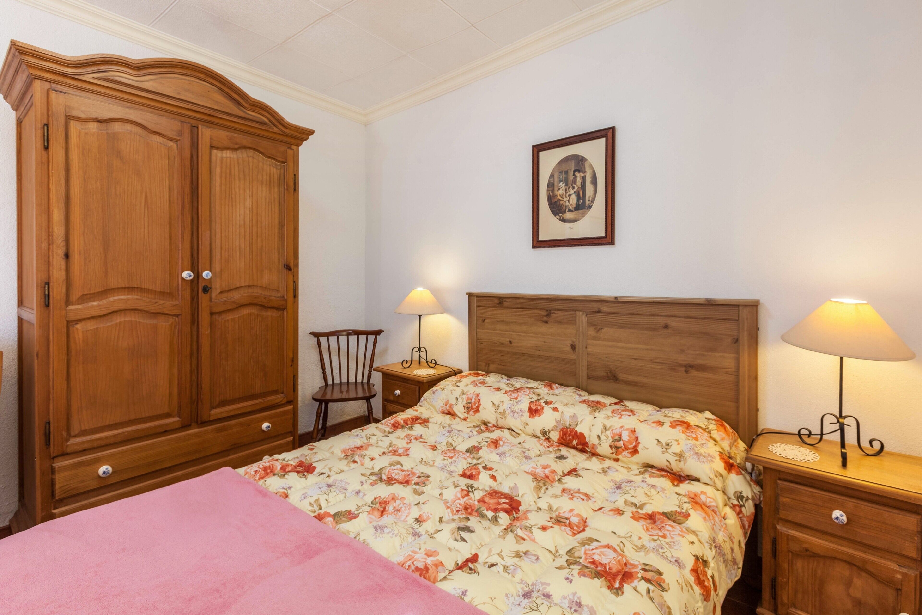 Quarto com guarda-roupa de madeira, cama com roupa de cama floral, duas mesas de cabeceira com abajures e uma cadeira.