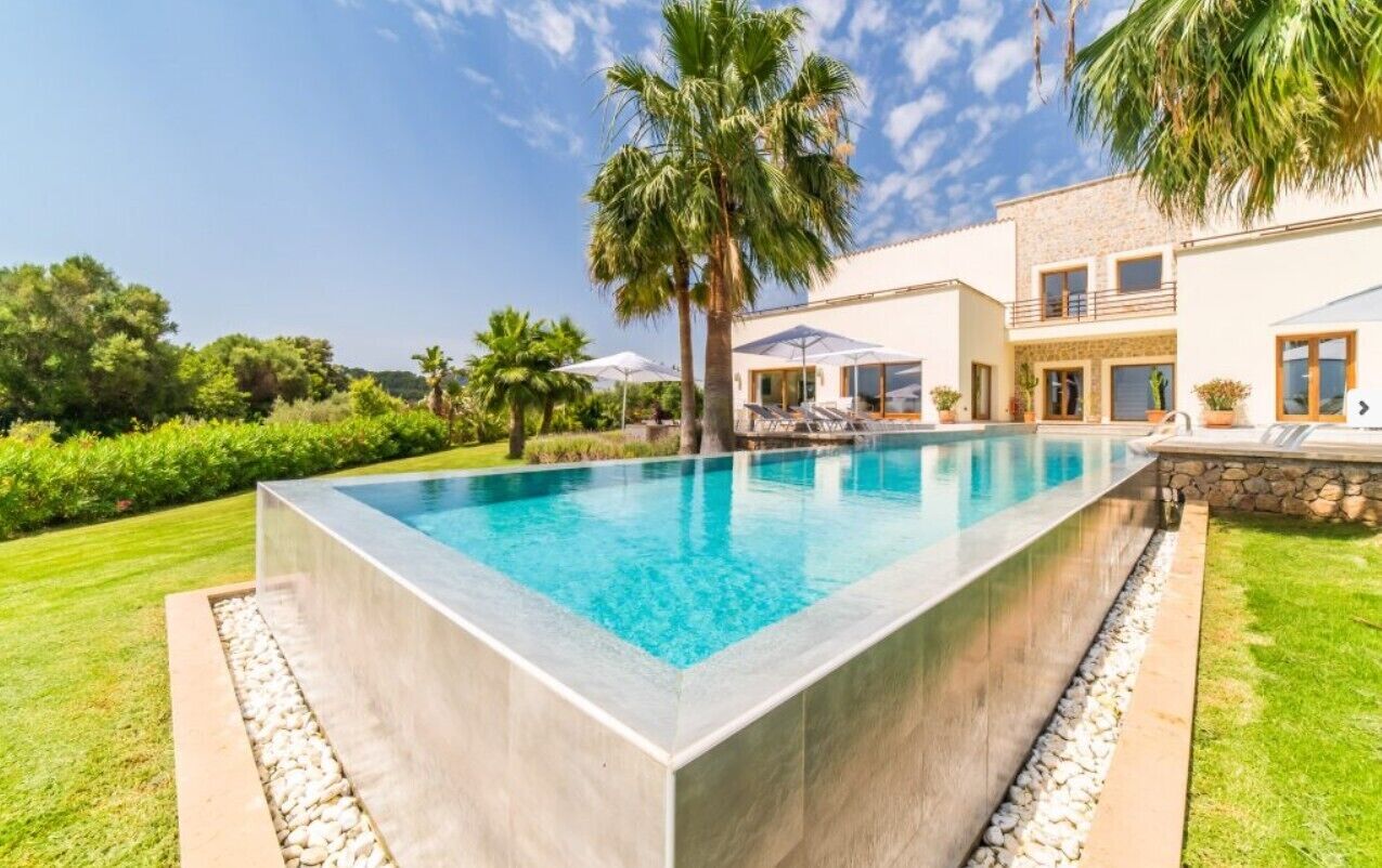 A rectangular swimming pool with a stone border, surrounded by a well-maintained lawn and palm trees.