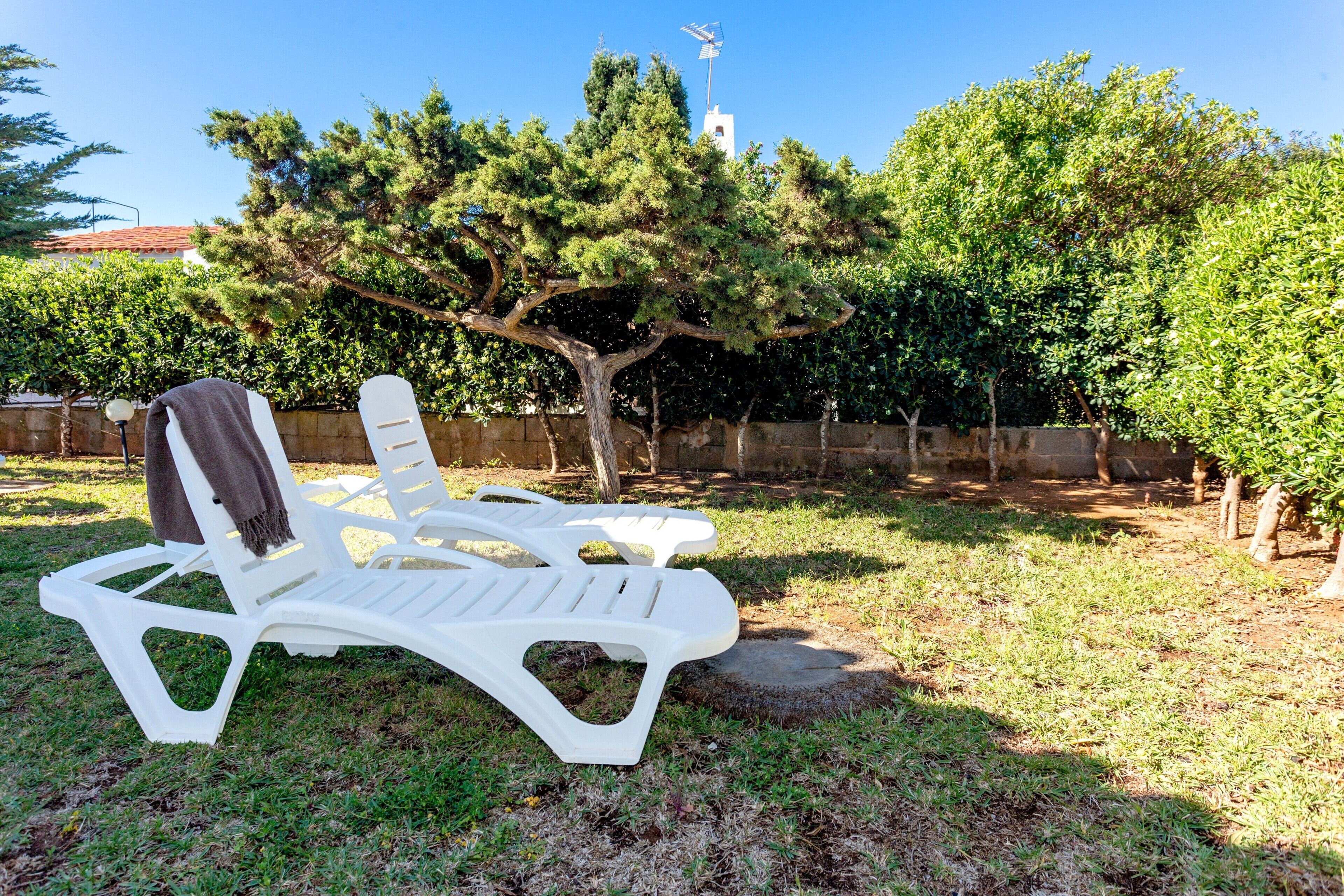 A white plastic lounge chair with a towel, a tree, and a stone wall in a garden.