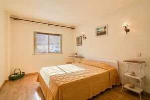 A bedroom with a bed, a window, a bedside table, a lamp, and a wall-mounted shelf.