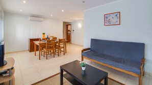 A living room with a sofa, a coffee table, a dining table with chairs, and an air conditioning unit on the wall.
