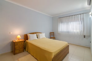 A bedroom with a bed, a nightstand with a lamp, a chair, and a window with curtains.