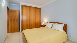 A bedroom with a bed, wooden wardrobe, and a nightstand with a lamp.