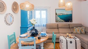 Et spiseområde med et rundt bord, stole og en vase med blomster. Der er en sofa og et billede på væggen.