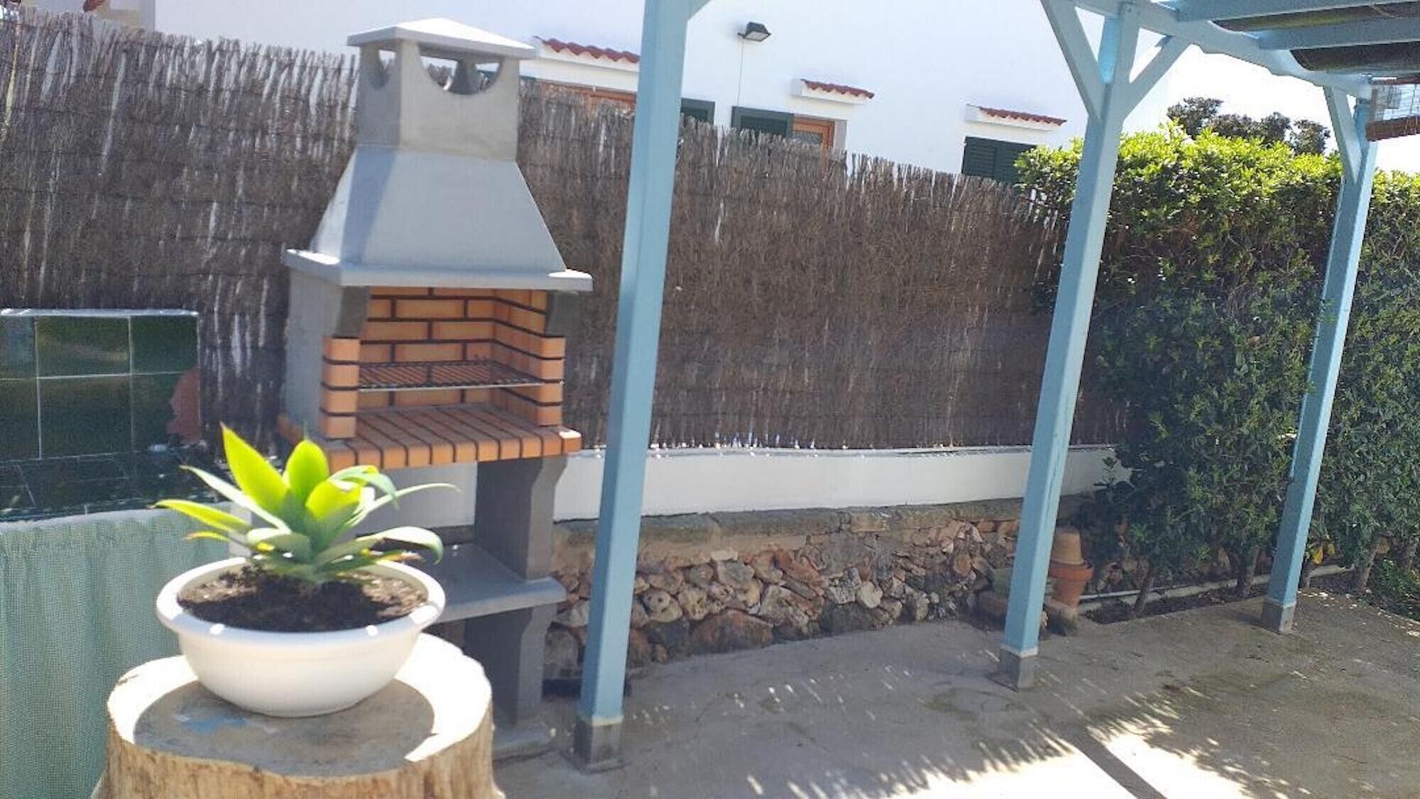 A brick outdoor oven under a blue metal structure, surrounded by a wooden fence and a white building in the background.