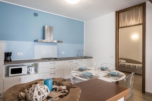 Cucina compatta con forno a microonde, bollitore e piano cottura a gas. C'è una zona pranzo con tavolo apparecchiato per due, sedie trasparenti e un divano sullo sfondo.