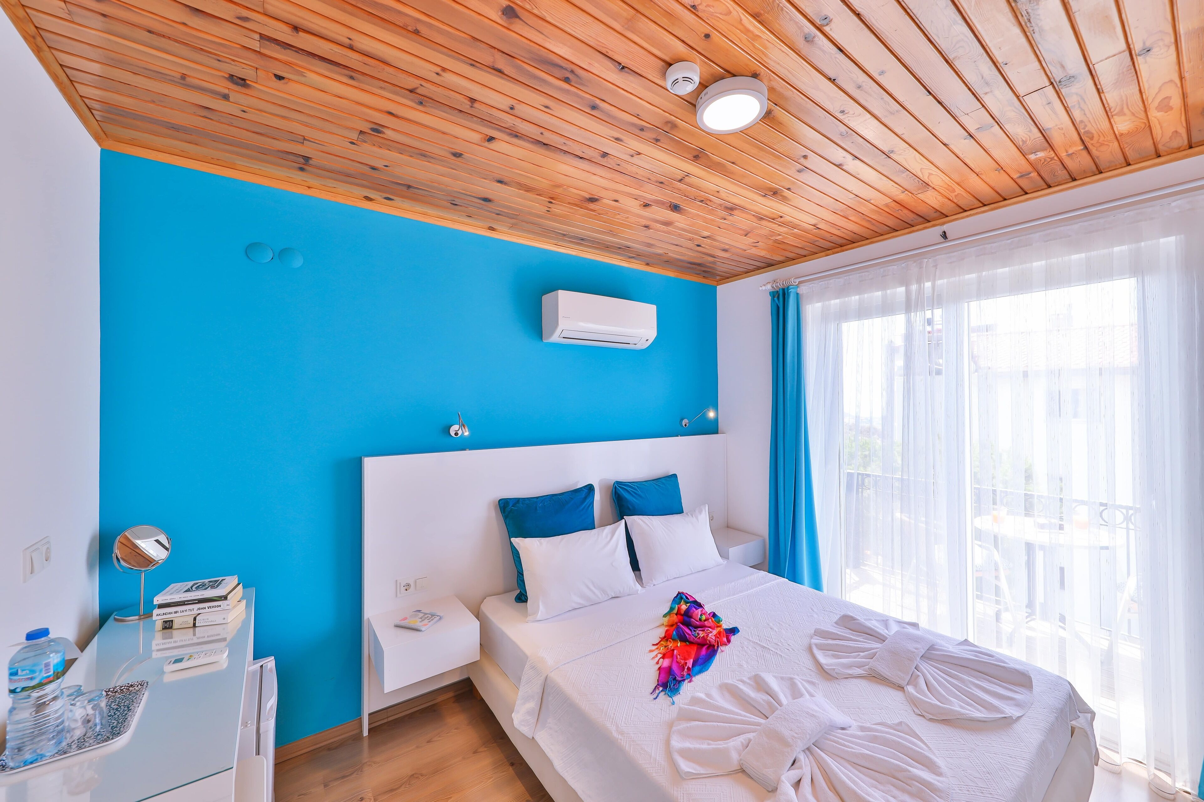 Ein Hotelzimmer mit Holzboden, einer leuchtend blauen Wand, einem Bett mit weißen Laken und einer bunten Decke, einem Nachttisch mit Lampe und einem Fenster mit durchsichtigen Vorhängen.