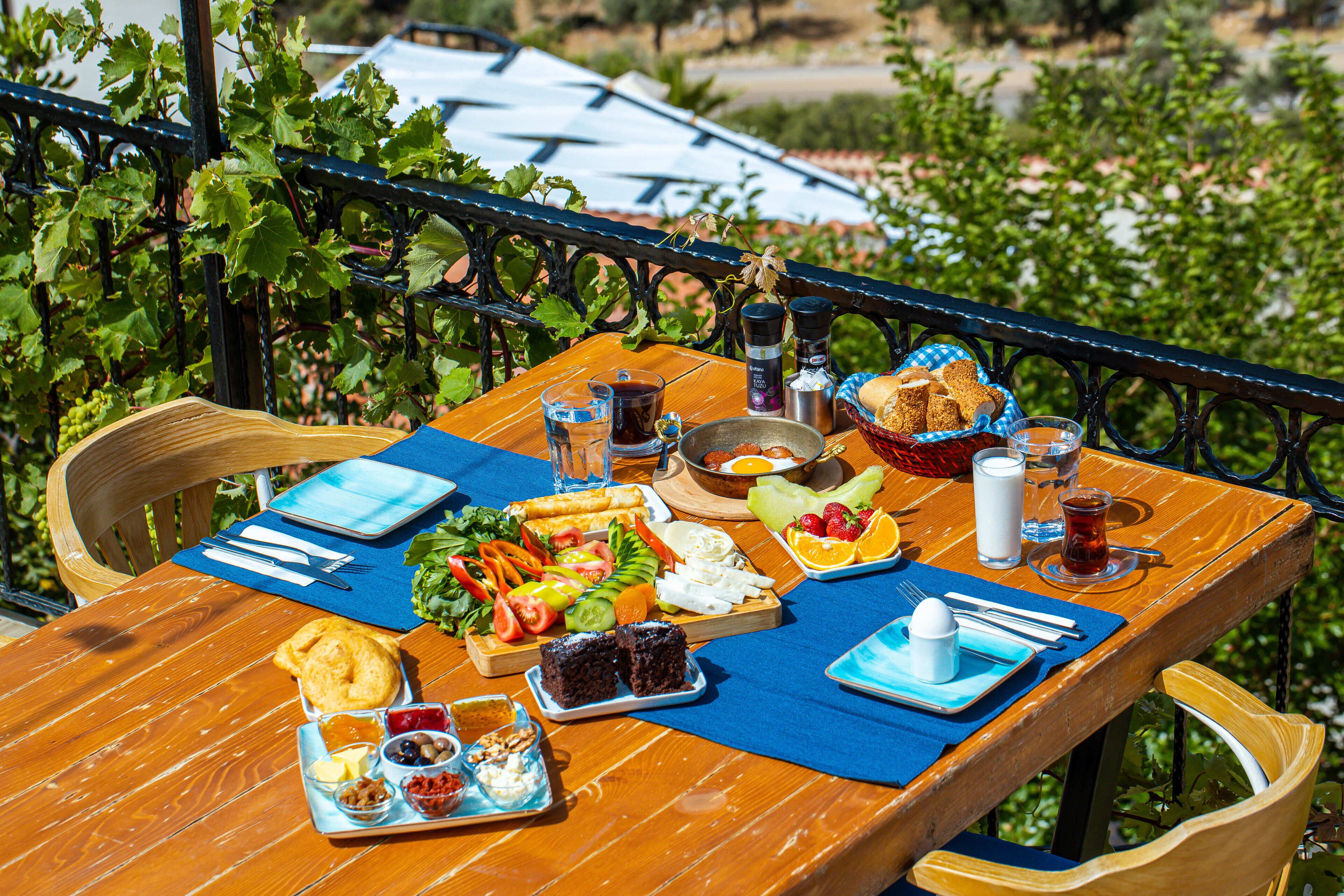 A wooden table set with a variety of food items, including bread, fruits, and a salad, on a terrace with a view of greenery and buildings in the background.