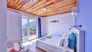 A bedroom with a wooden ceiling, a bed with pillows, a bedside table with a book, a window with a view of buildings, and a wall-mounted light.