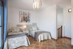 A bedroom with two beds, a nightstand, a lamp, a painting of bottles, and a staircase.