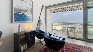 Un salon moderne avec une grande fenêtre, un tableau représentant un voilier, une bibliothèque, un fauteuil bleu et une petite table.