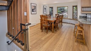 A spacious dining area with a wooden table and chairs, a staircase with a metal railing, and large windows offering a view of the outdoors. - Eiger 9 3 Bedroom Condo by RedAwning (Vail)