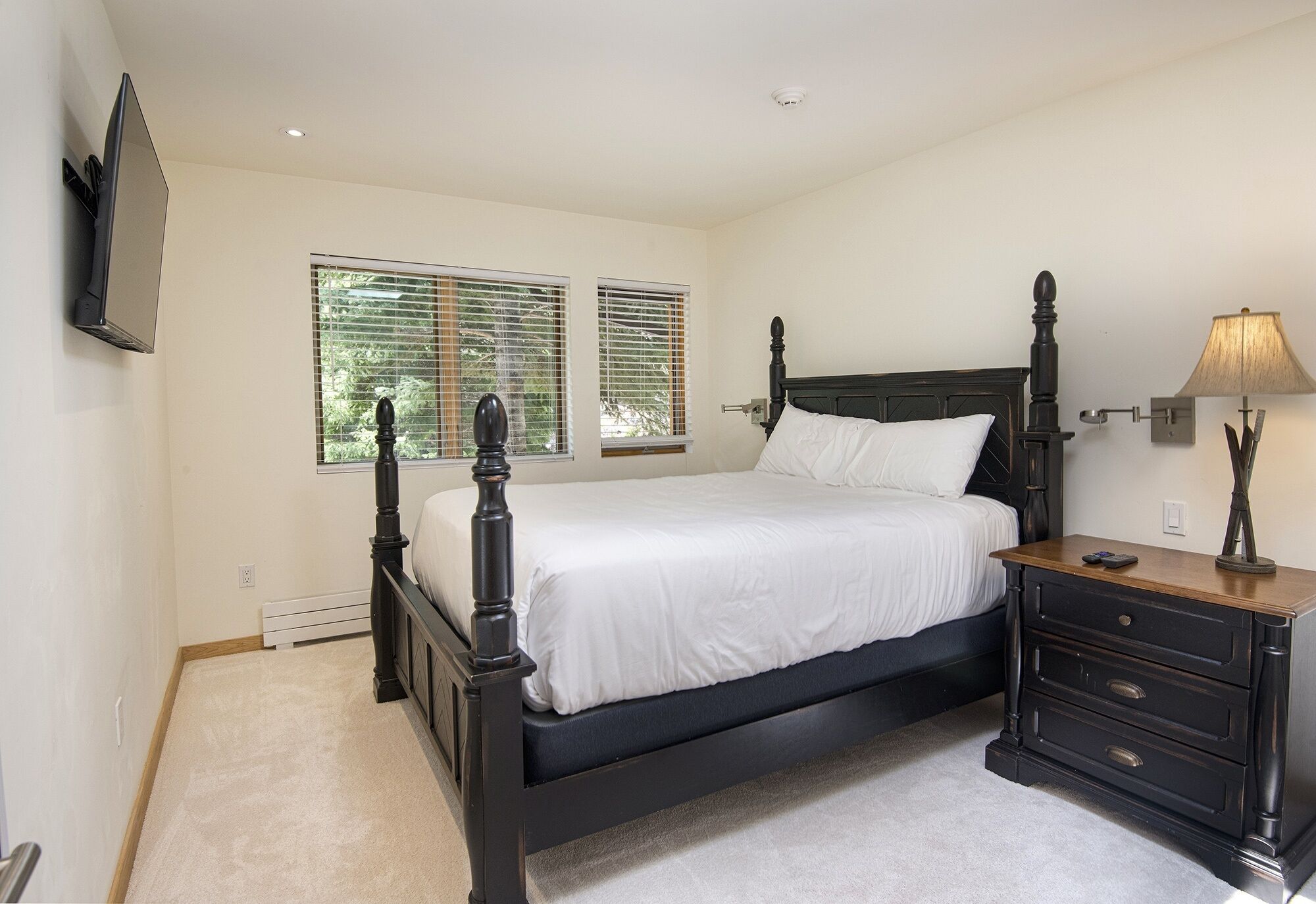 A bedroom with a four-poster bed, a nightstand with drawers, a lamp, and a TV mounted on the wall.