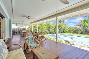 Amplo espaço ao ar livre com deck de madeira, móveis de vime e vista para uma piscina e vegetação exuberante.