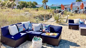 Ein Strandloungebereich mit Korbmöbeln, einer Kühlbox mit Getränken und Blick auf Strand und Hotelgebäude.
