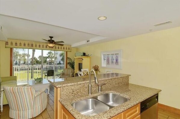 Cocina con encimeras de granito, pileta de acero inoxidable y muebles de madera. Hay un comedor con una silla a rayas y un ventilador de techo.