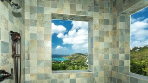 Un baño con vistas a un paisaje costero a través de una ventana, paredes con azulejos y un cabezal de ducha.