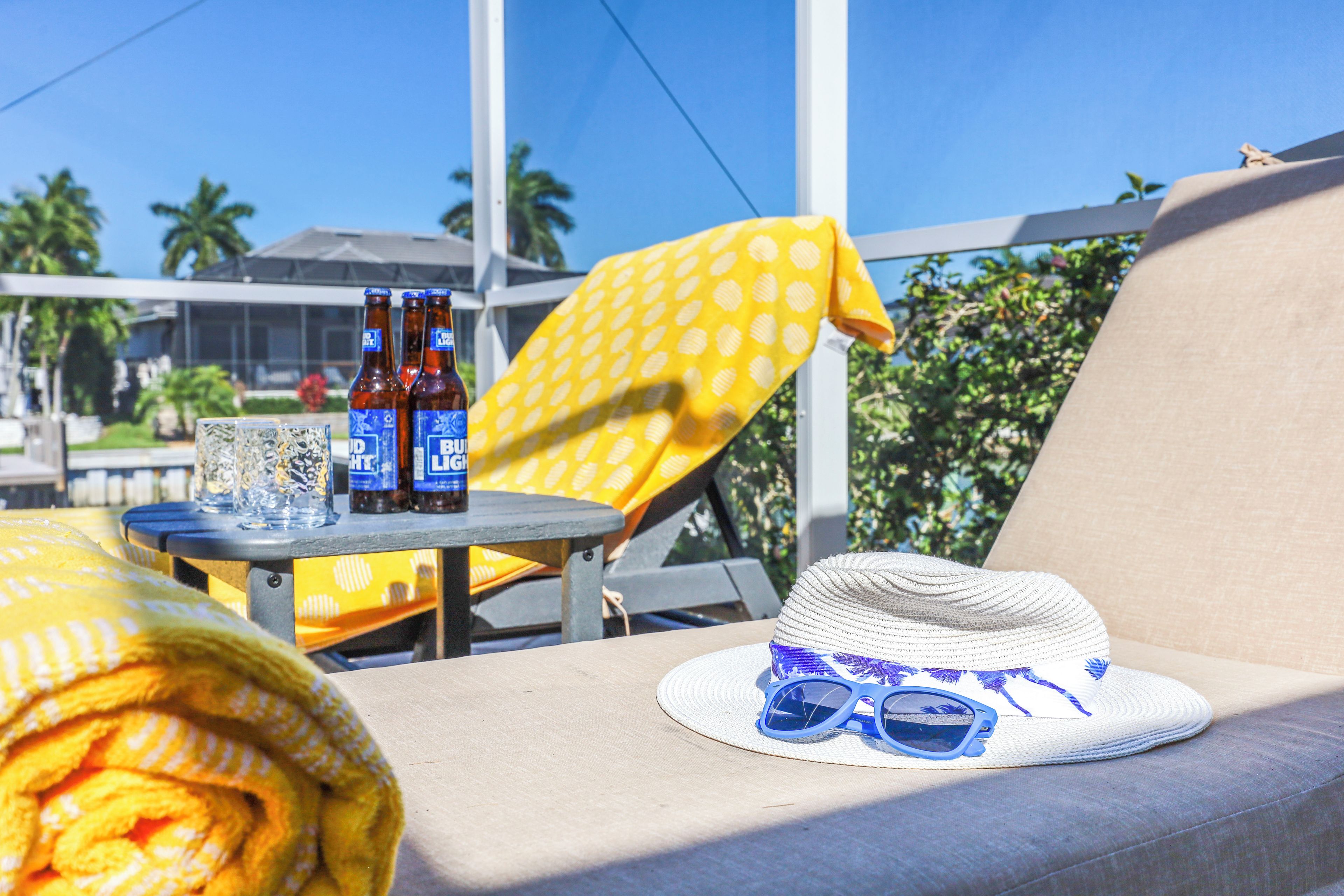 Una tumbona con sombrero y gafas de sol, una mesa con botellas y vasos, y una toalla amarilla.