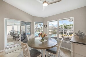 A dining area with a round table, chairs, a vase of flowers, and large windows offering a view of a pool and outdoor area.