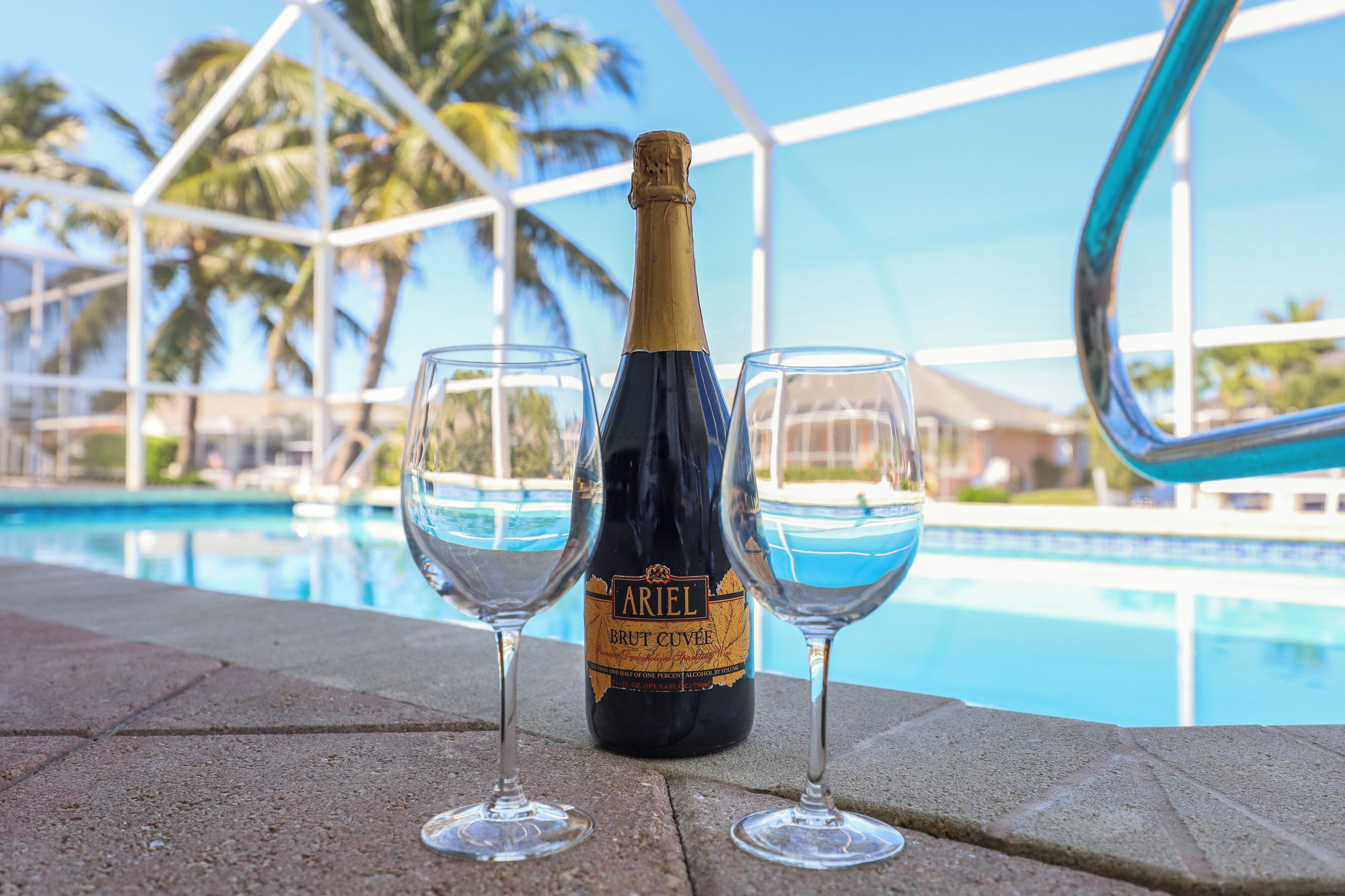 A bottle of Ariel Brut Cuvee champagne and two glasses on a poolside with a clear view of the pool and palm trees.