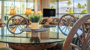 Una mesa redonda de vidrio con dos sillas, una planta en maceta y un televisor de pantalla plana en una habitación con ventanas amplias y vista a palmeras.