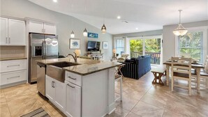 A spacious kitchen with a central island, white cabinetry, stainless steel appliances, pendant lighting, and a view to an outdoor seating area.