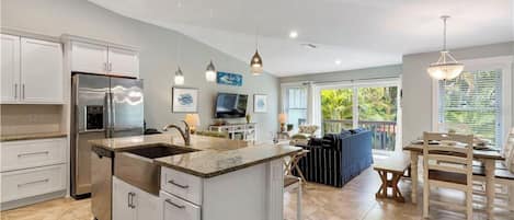 A spacious kitchen with a central island, white cabinetry, stainless steel appliances, pendant lighting, and a view to an outdoor seating area.