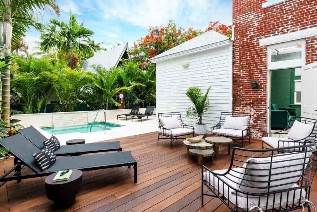 A poolside area with lounge chairs and a small table, surrounded by lush greenery and a brick building.