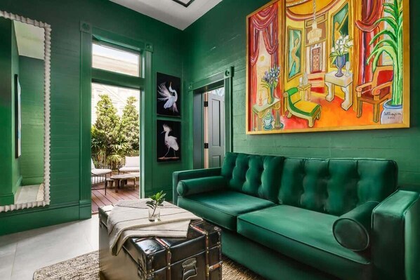 A room with a green wall, a green sofa, a painting, and a window with a view of trees.