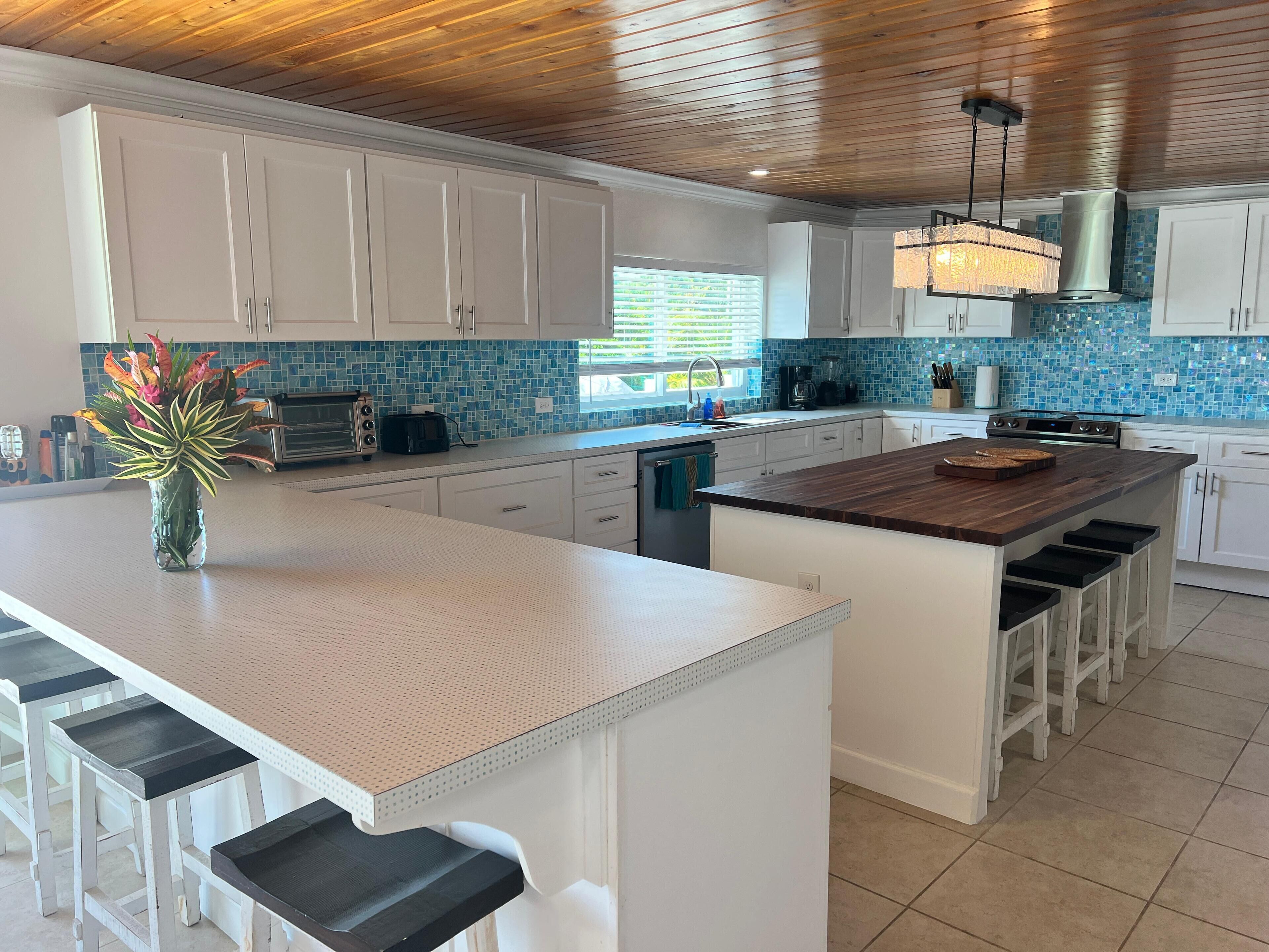 A modern kitchen with white cabinets, a wooden ceiling, a blue-tiled backsplash, a central island with stools, and a hanging light fixture.
