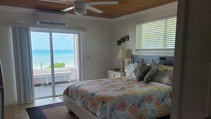 A bedroom with a bed, a nightstand, a lamp, a ceiling fan, a window with blinds, and a view of the ocean.