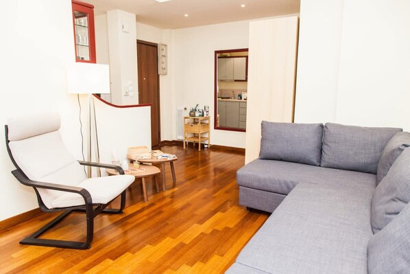 A living room with a grey sofa, a white chair, a small wooden table, and a lamp. - Centrally Located Studio In Athens (Athens)