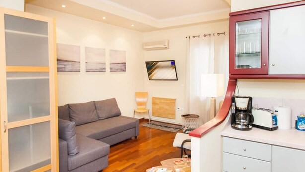 A compact living space with a grey sofa, a small round table with books, a coffee machine, and a cabinet with glass doors. - Centrally Located Studio In Athens (Athens)
