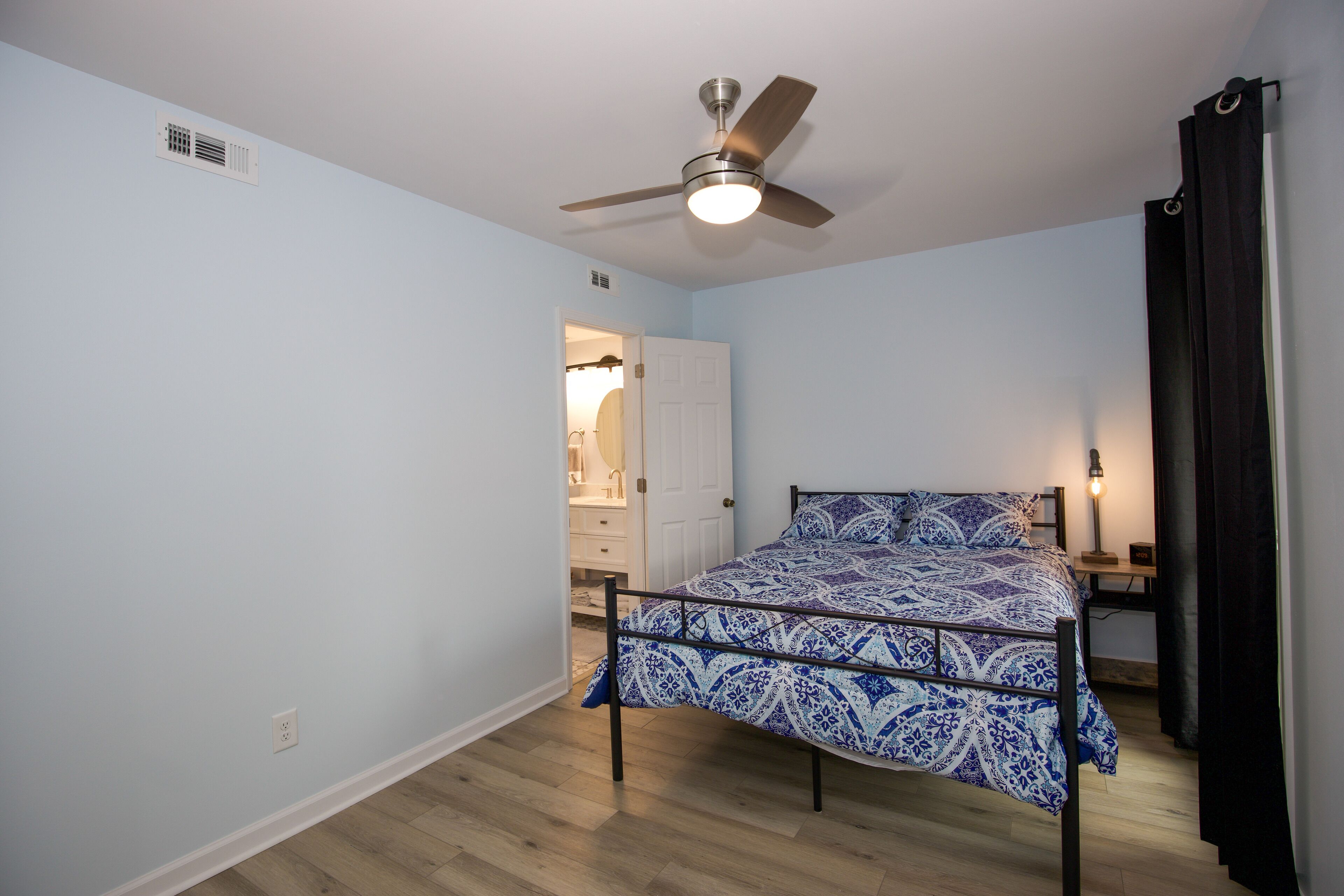 A bedroom with a bed, a ceiling fan, a nightstand, a lamp, and a door leading to a bathroom.