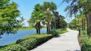 A paved pathway alongside a body of water with palm trees and greenery. - Resort Condo 3BR w Balcony, Free Parking near OCCC (Orlando)