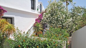 Un edificio blanco con balcón, rodeado de diversas plantas florecientes y una palmera.