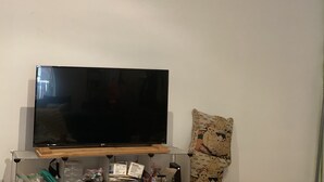 Litteänäyttöinen televisio on sijoitettu puiseen jalustaan valkoista seinää vasten, ja oikealla puolella on kuvioitu koristetyyny.
