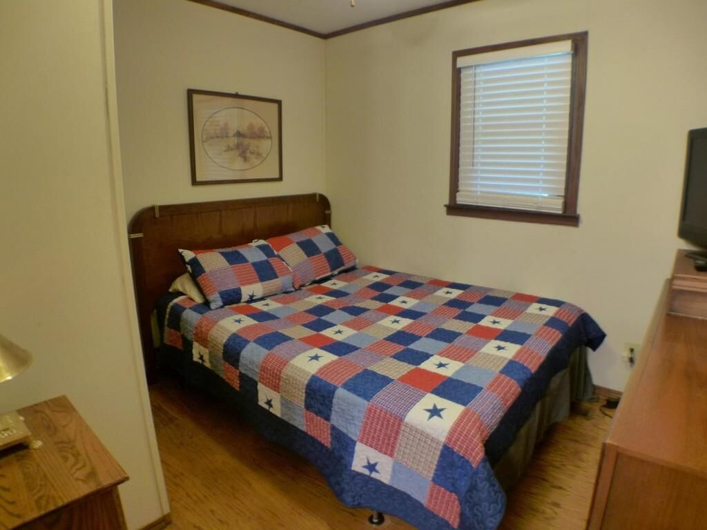 Un dormitorio con cama, un cuadro en la pared, una ventana con persianas y un televisor sobre un mueble.