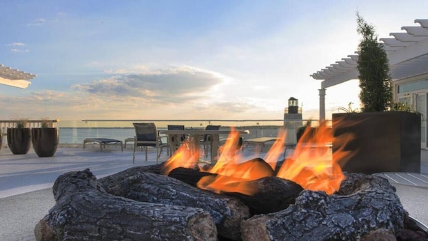 A fire pit with logs and flames, surrounded by outdoor seating and a view of the ocean.