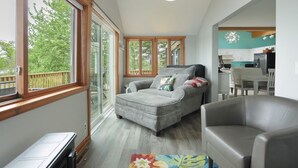 A spacious living area with a grey sofa, a grey armchair, and a colorful rug. There is a large window providing a view of trees outside. - Enchanting, Romantic Lakefront Cottage, Close to Frankenmuth (Millington)