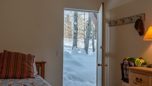 Une chambre à coucher avec un lit, un banc en bois, une porte donnant sur un espace extérieur enneigé, un porte-manteau et une lampe.