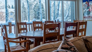 Un coin repas avec une table et des chaises en bois, de grandes fenêtres donnant sur des arbres couverts de neige et une maison.