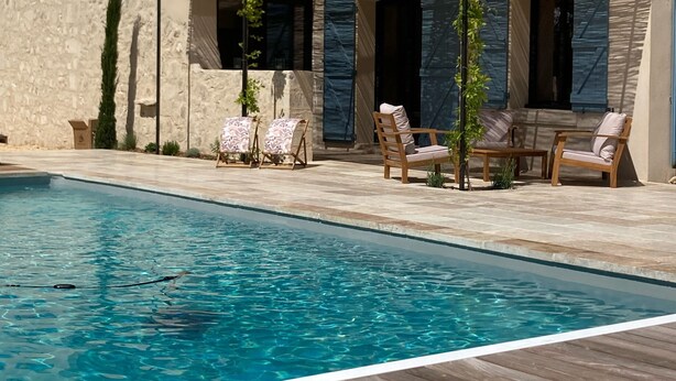 A swimming pool with a wooden deck and lounge chairs, a stone building with a tiled roof, and a clear blue sky.
