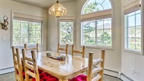 Un comedor con una mesa y sillas de madera, ventanas amplias con persianas y una lámpara colgante.