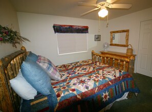 A bedroom with a wooden bed, a quilt, a bedside table with a fan, a mirror, and a window with a view of the outdoors.